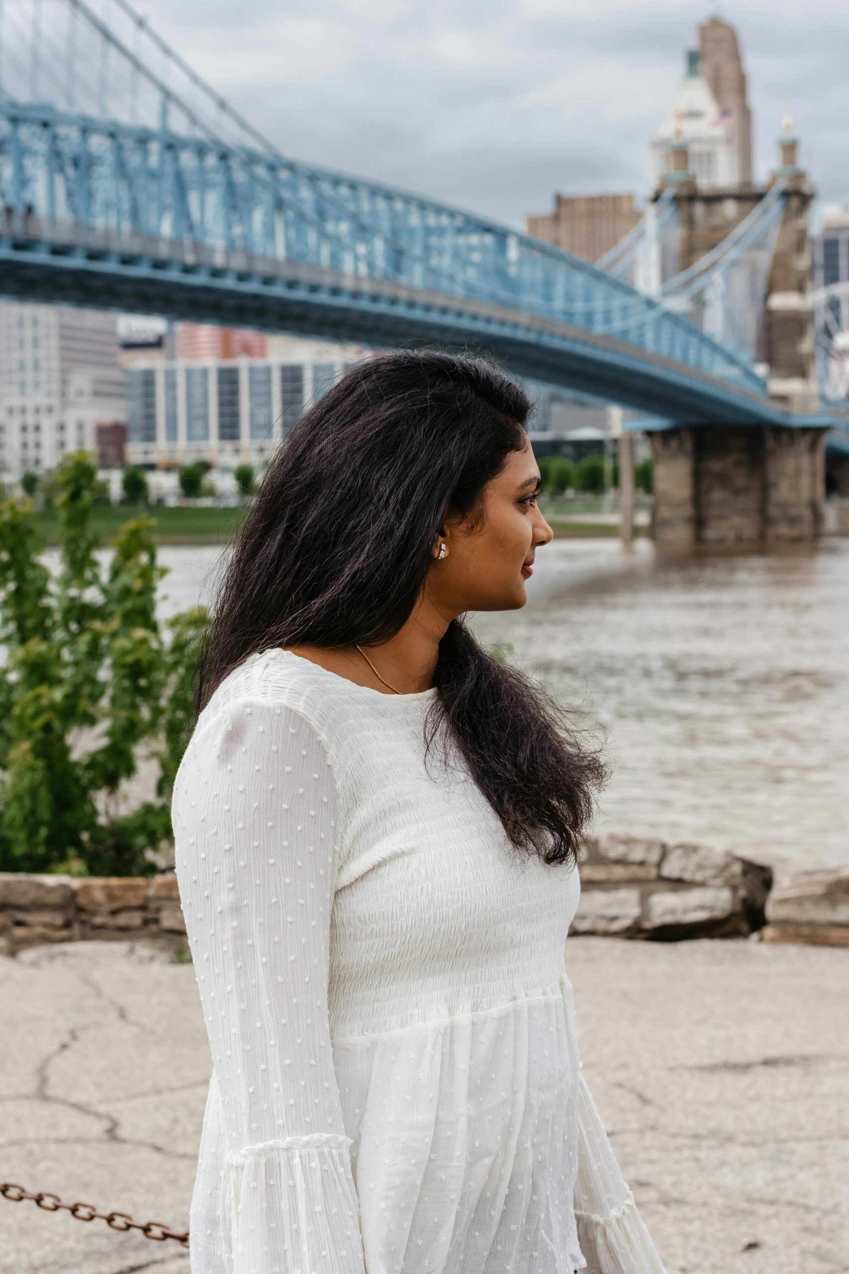 A woman in a white dress stands by the river with the John A. Roebling Suspension Bridge in the background.